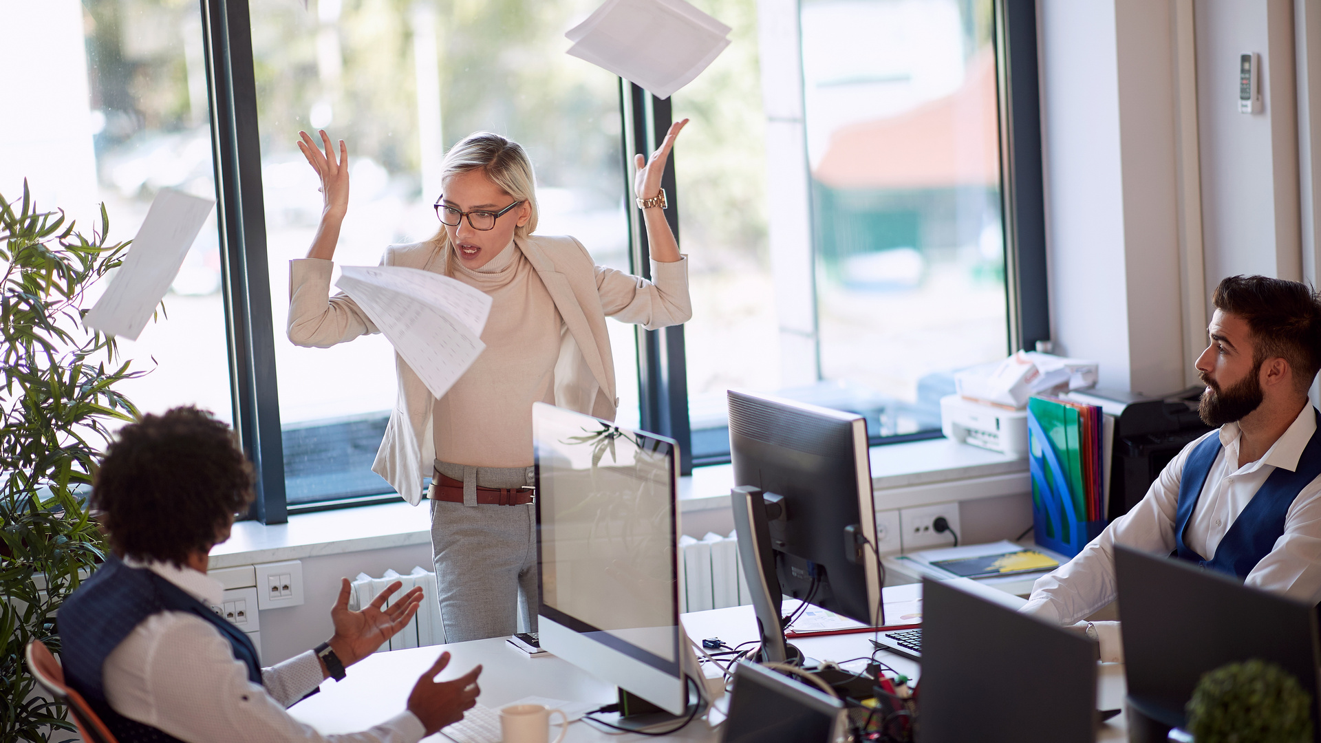 Kollegin regt sich im Büro auf und wirft Papier um sich, Mitarbeiter sitzen vor PCs und diskutieren mit